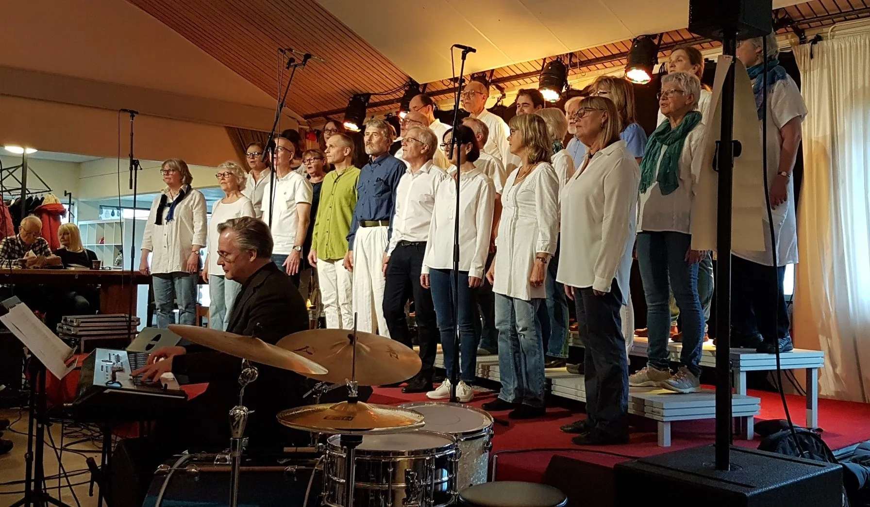 Kören i vita och gröna kläder på scenen på Kulturoasen, med musiker i förgrunden.